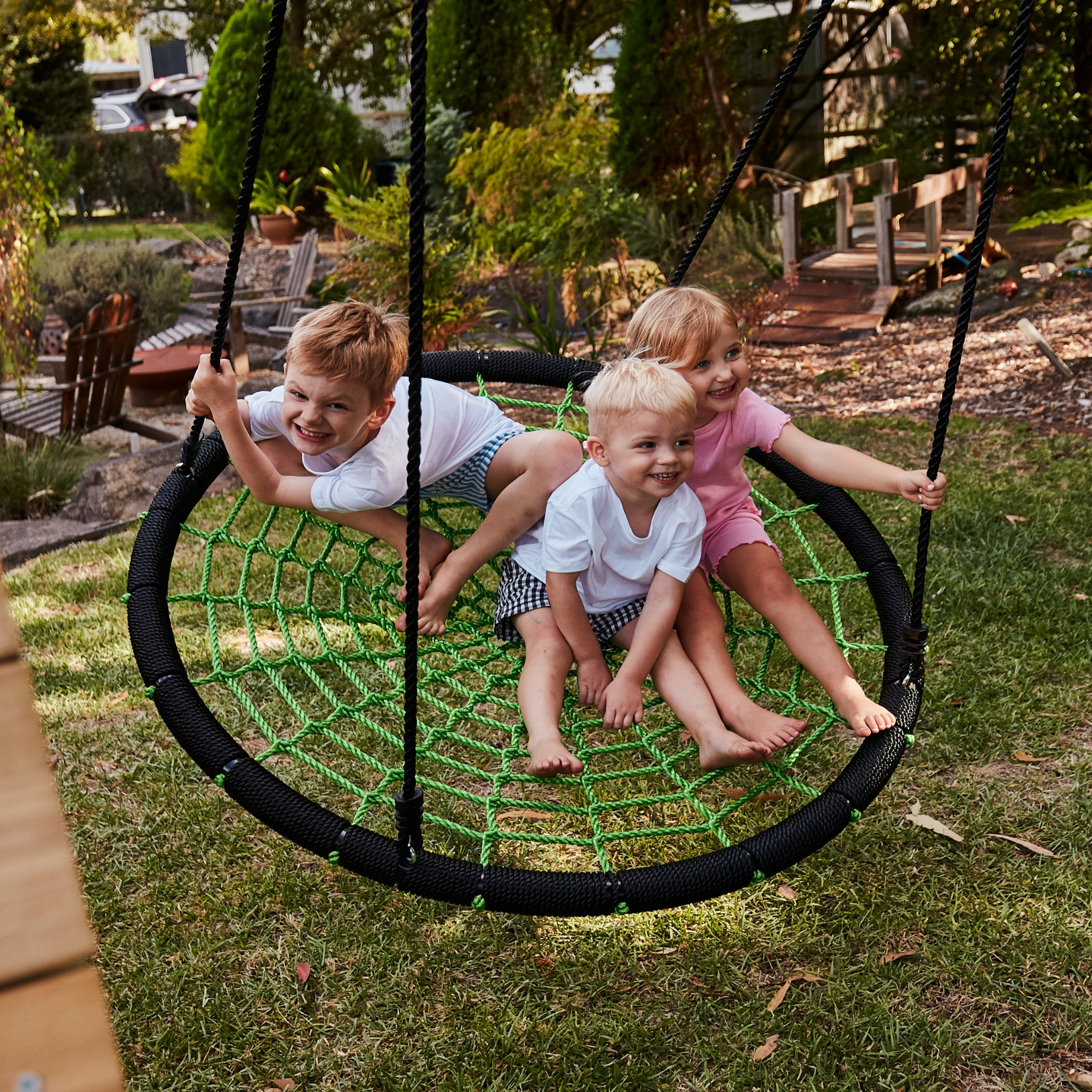 Three children on a round swing set in a backyard with trees and a wooden deck.