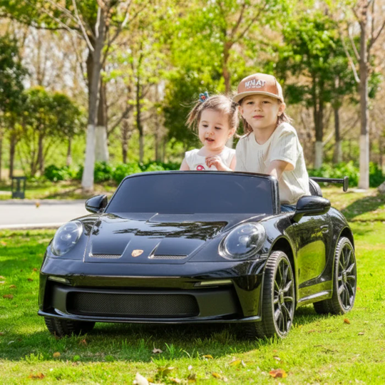 Two children sitting in a black toy car on a grassy area with trees in the background