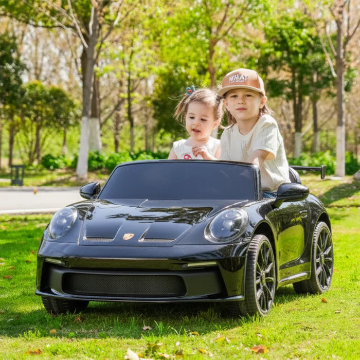 Two children sitting in a black toy car on grass with trees in the background