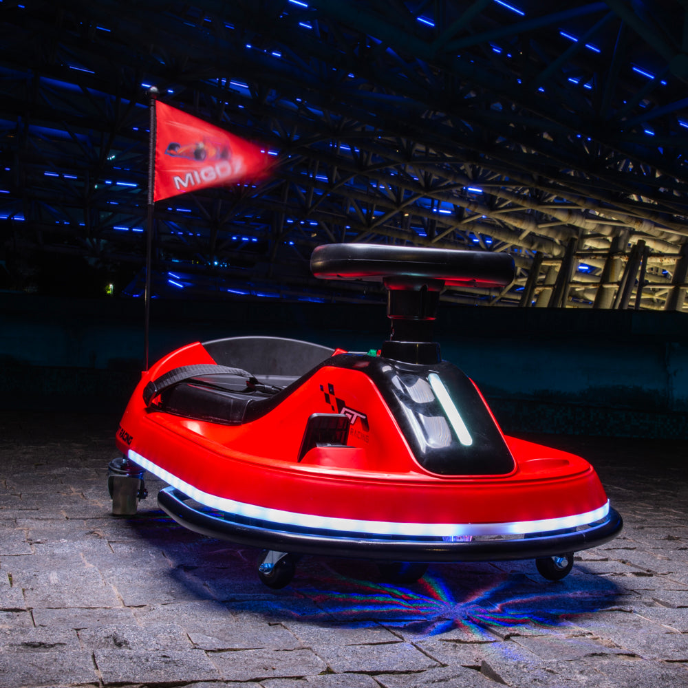 A red electric bumper car with a black seat and steering wheel, featuring integrated white LED strip lights along the bottom trim and a red flag on the rear with the brand name 'MIOO'. The vehicle is parked on a stone floor inside a dark, industrial hall with blue ambient lighting.