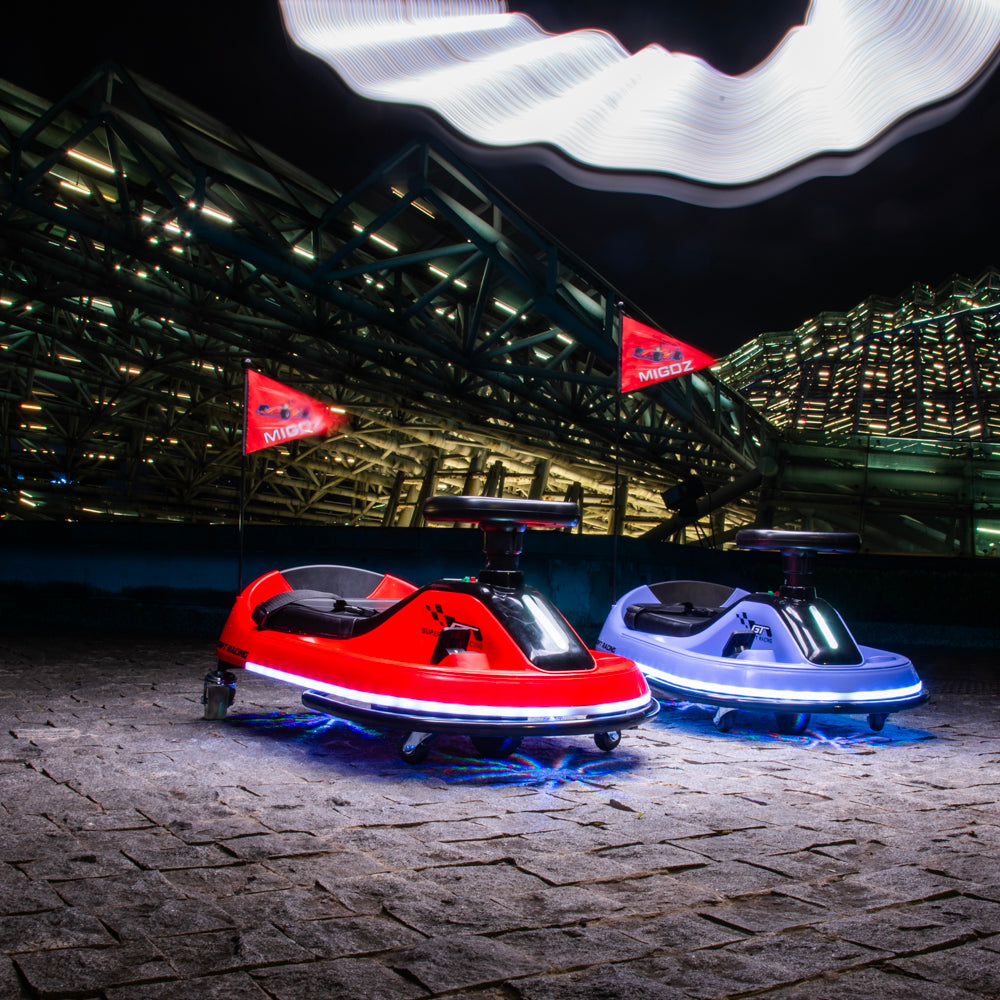 Two small electric bumper cars, one red and one light blue, parked on a stone surface in front of a large illuminated industrial structure at night. Each car has a small red flag with the brand name 'MOBIL' visible.