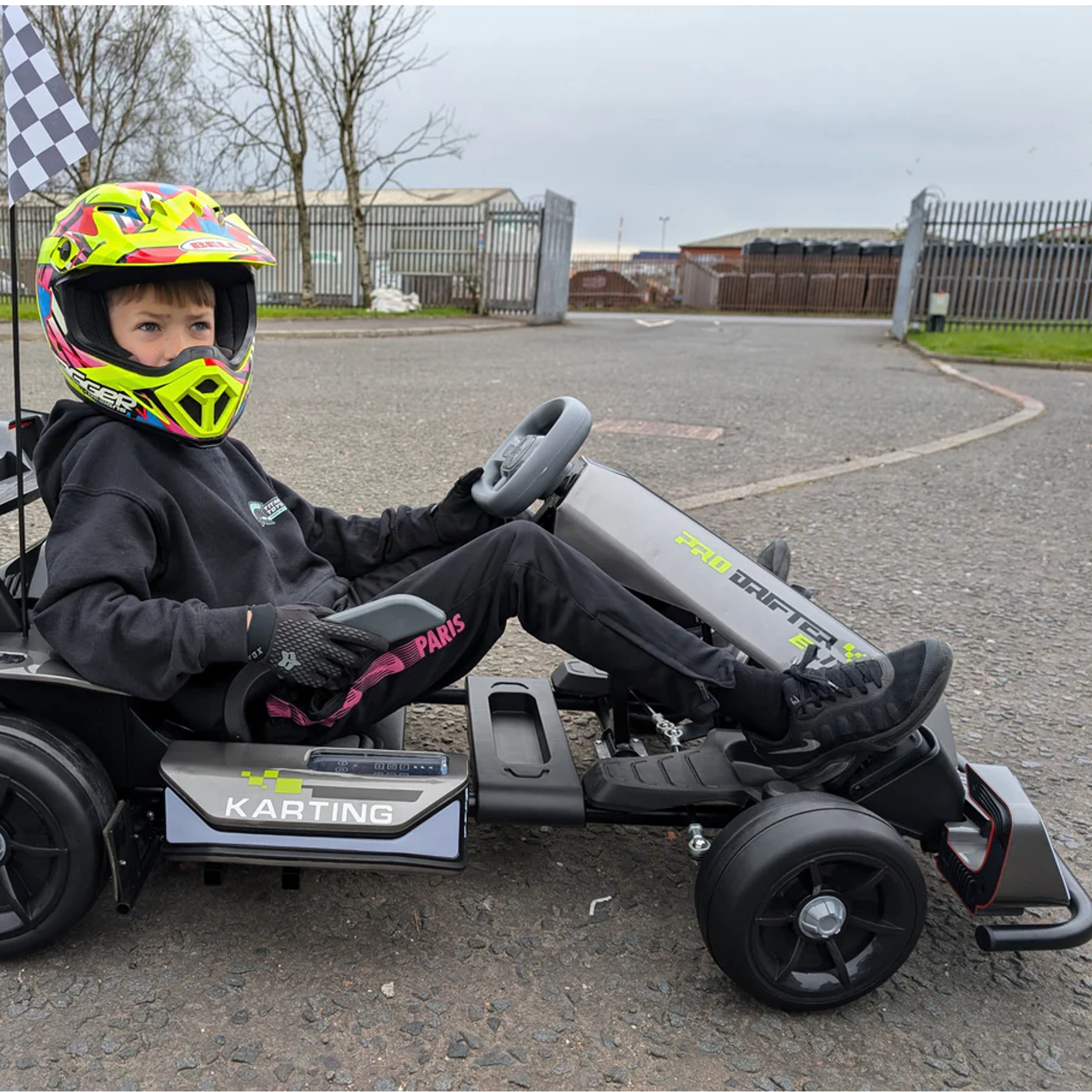 Child sitting in a go-kart wearing a helmet with a checkered flag in the background.