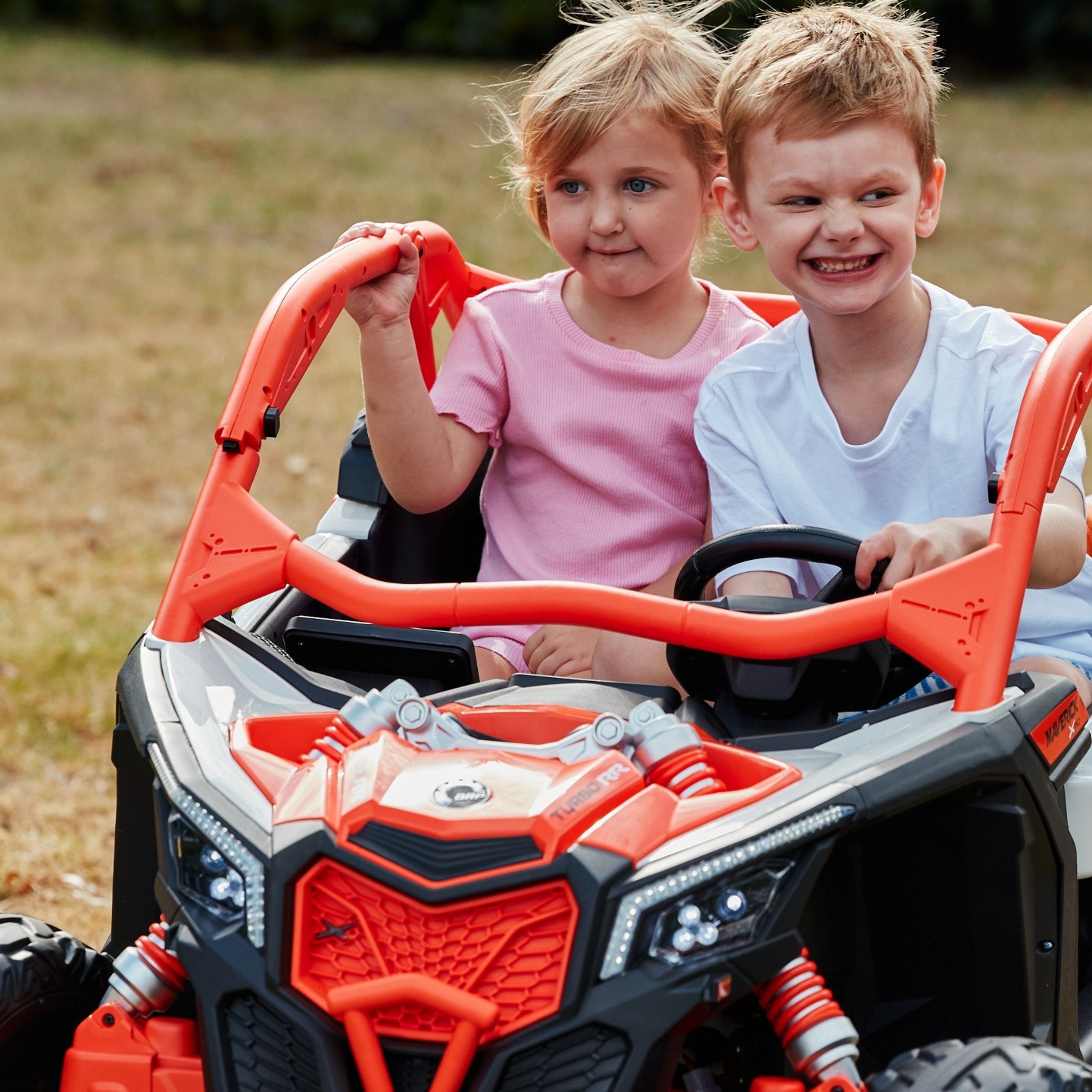 Two children sitting in a toy off-road vehicle outdoors.