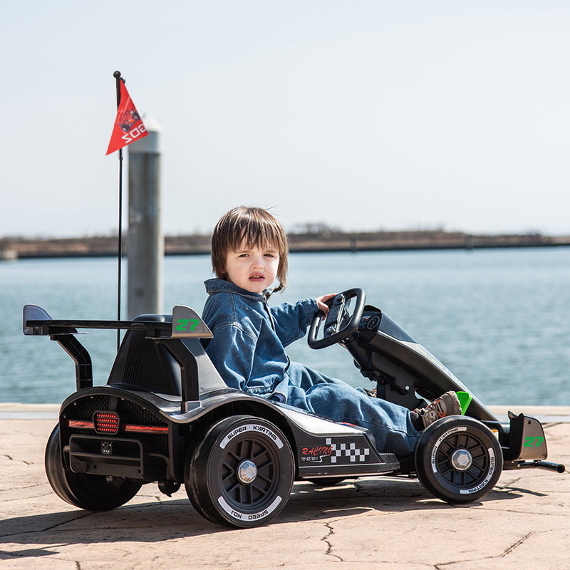 A young child wearing a blue denim jacket and jeans sits in a black go-kart on a paved waterfront area, with water and a distant pier visible in the background. The go-kart has green and white decals, including a checkered pattern near the front right side.