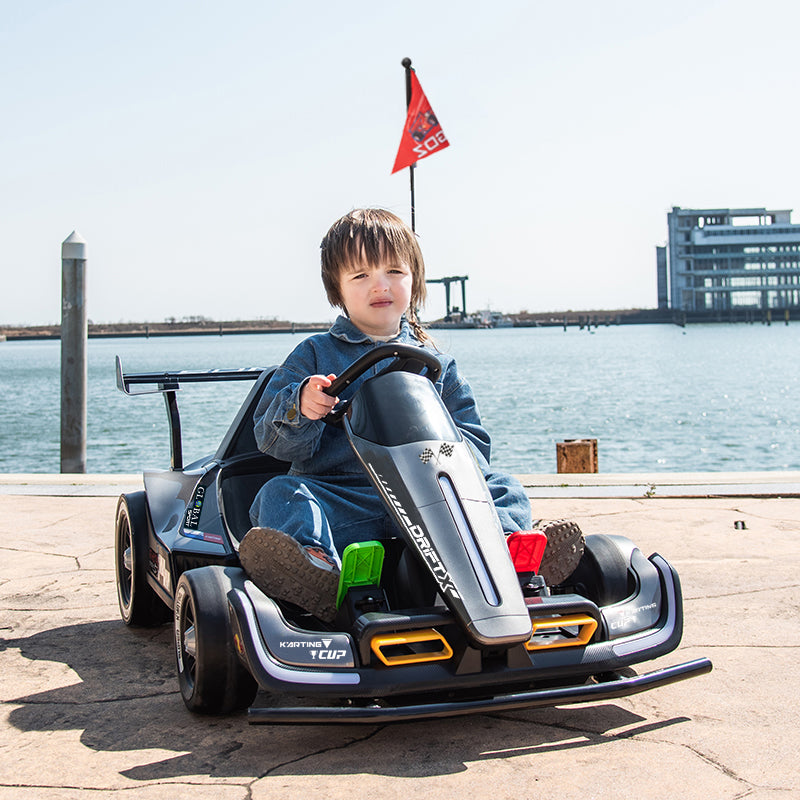 A young boy wearing a denim jacket and jeans sits in a black and gray toy go-kart on a paved waterfront promenade, with water, a pier, and a building visible in the background.