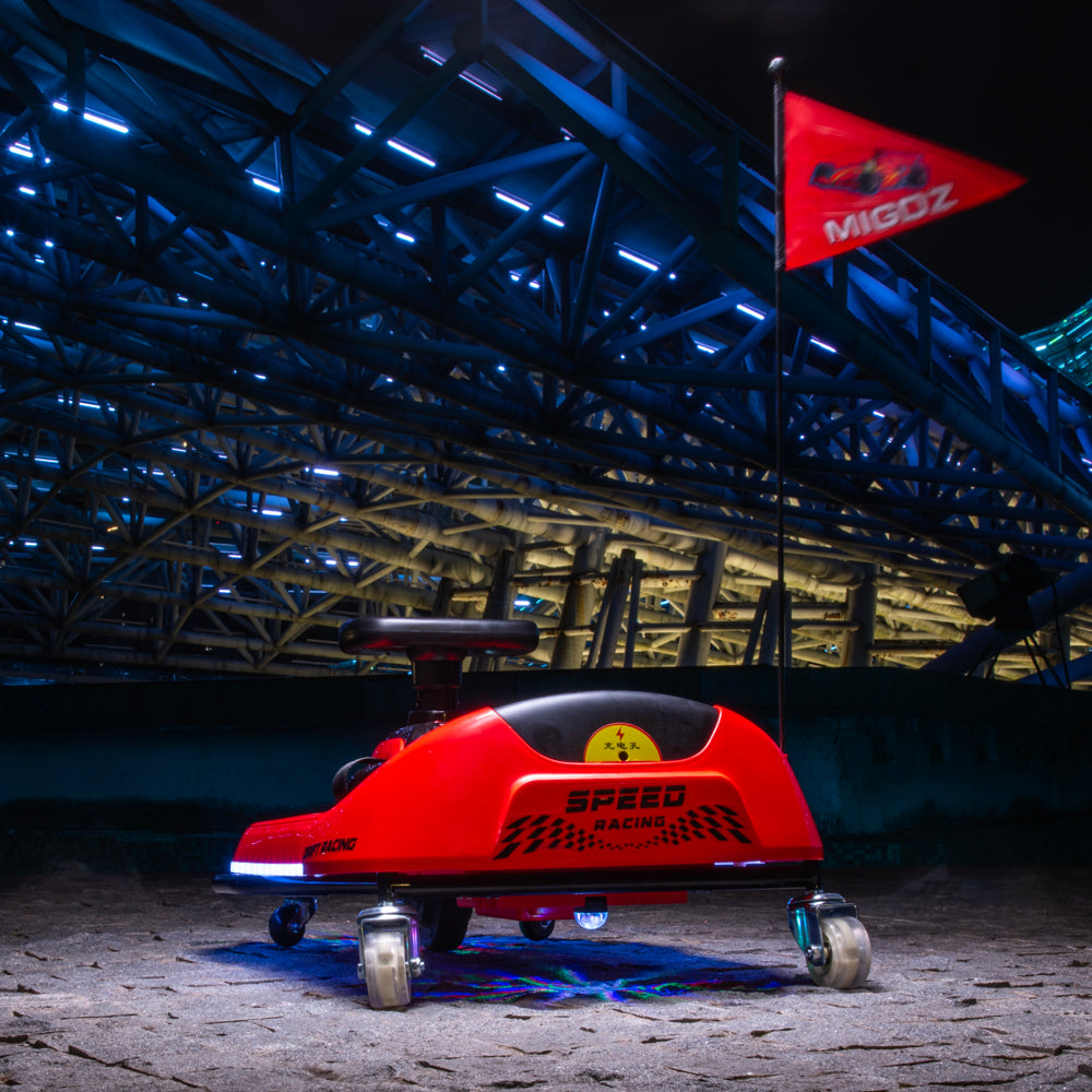 A red MigoZ electric speed racing cart with a black seat and wheels, positioned in front of a large illuminated stadium structure at night with a red MigoZ flag in the background.