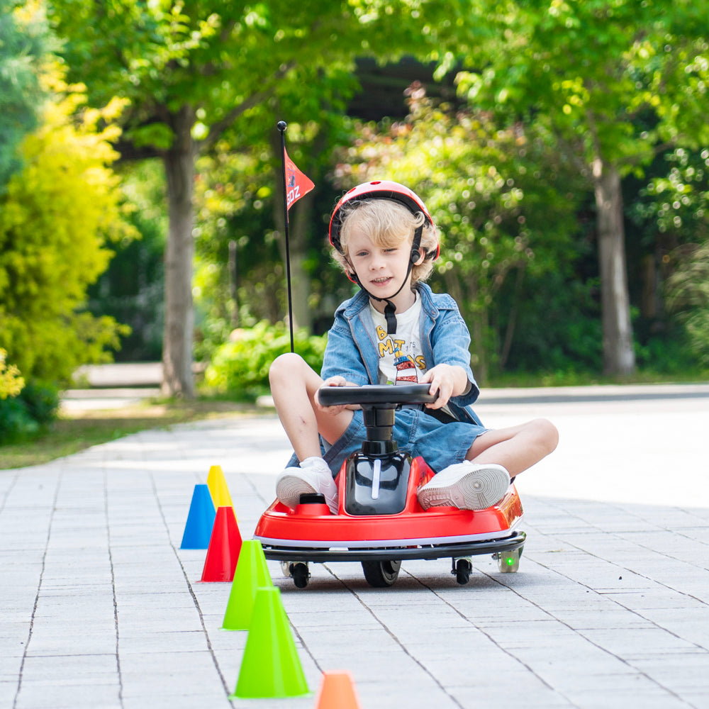 A young child wearing a red helmet sits on a red pedal toy car, driving it through a line of colorful cones on a paved path with a blurred green park background.