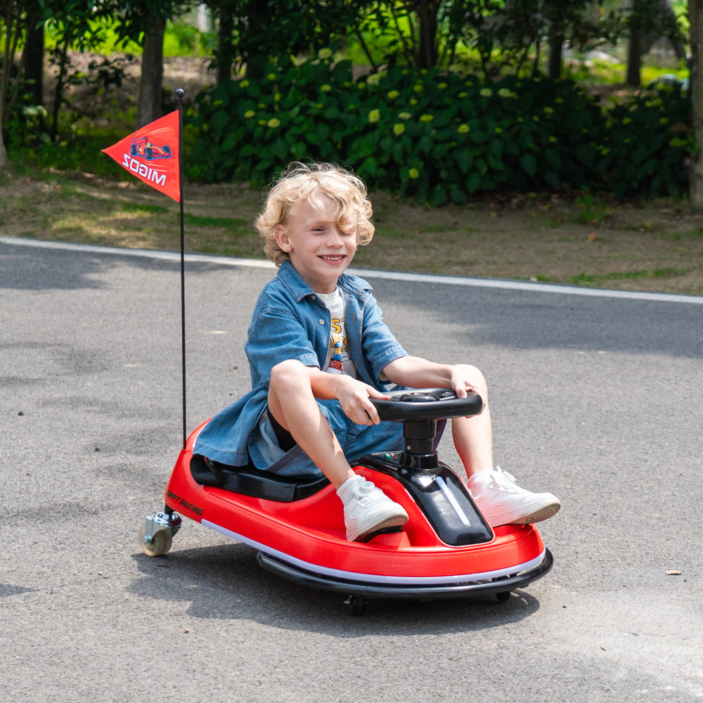 A young child with curly blonde hair sits smiling on a red go-kart toy outdoors on a paved road, with green trees and bushes in the background. The go-kart has a small red flag attached to the back with the brand name 'Mikomo' visible.