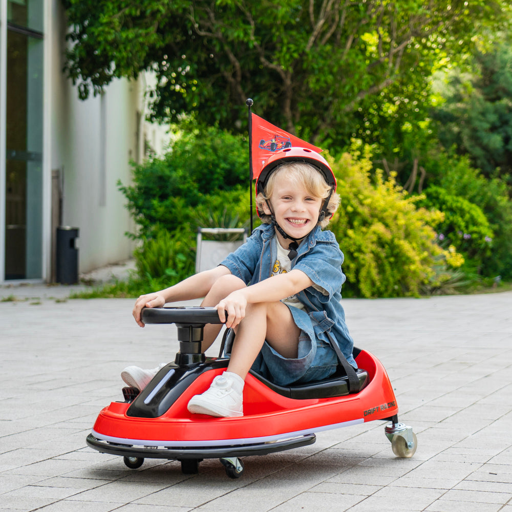 A young child wearing a red helmet and denim outfit sits smiling in a red pedal toy car on a paved outdoor area with green trees in the background.