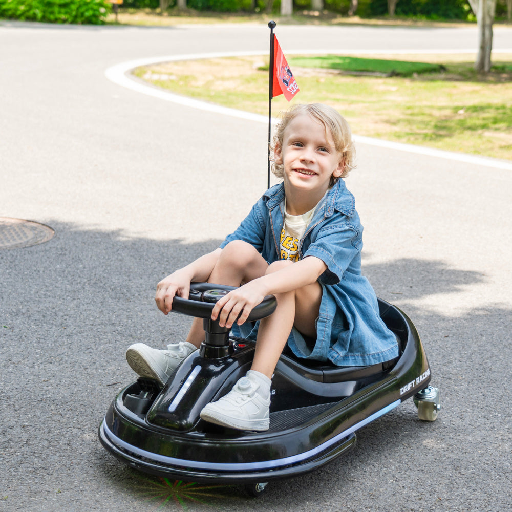 A young child wearing a denim shirt and white sneakers sits on a black plastic ride-on toy car with a small flag on a paved outdoor path.