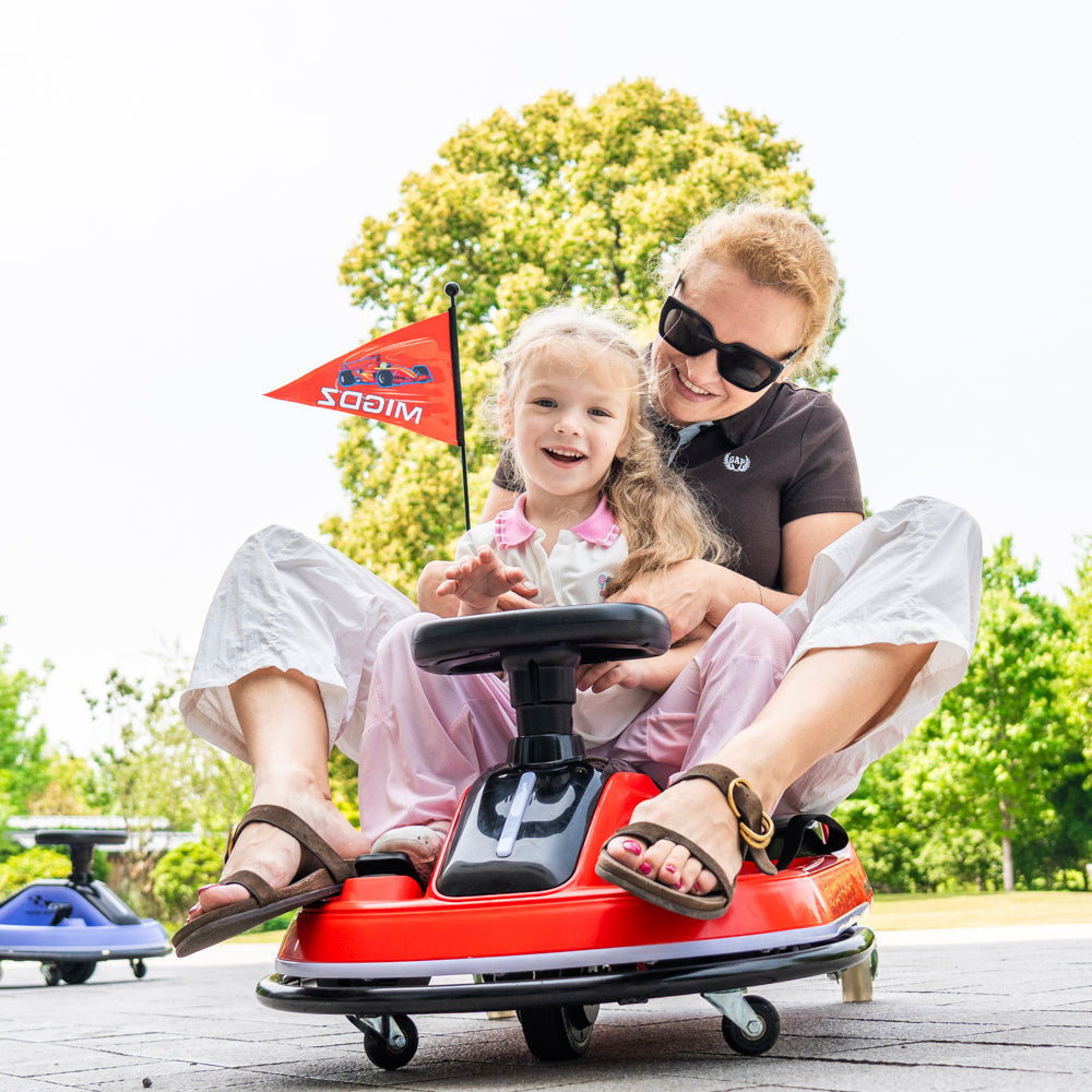A woman and a young girl enjoy a red SODIM brand kiddie wheeled car outdoors on a paved path with green trees in the background.