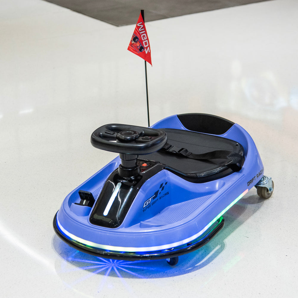 A blue and black electric toy car with a small red 'NINJA' flag on a pole, photographed on a clean white floor with a reflective surface.