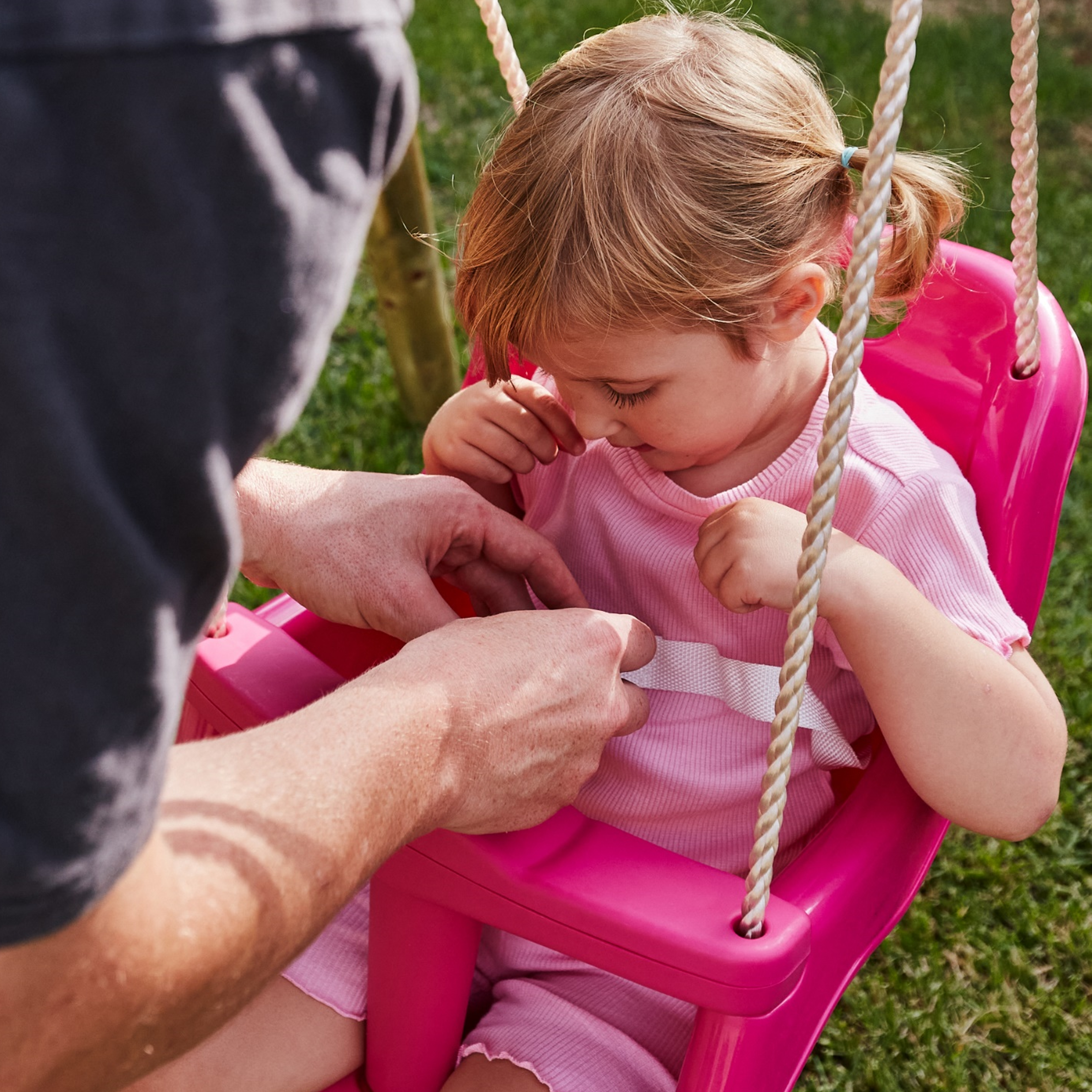 Man helping a young girl sit in a pink swing on a grassy area