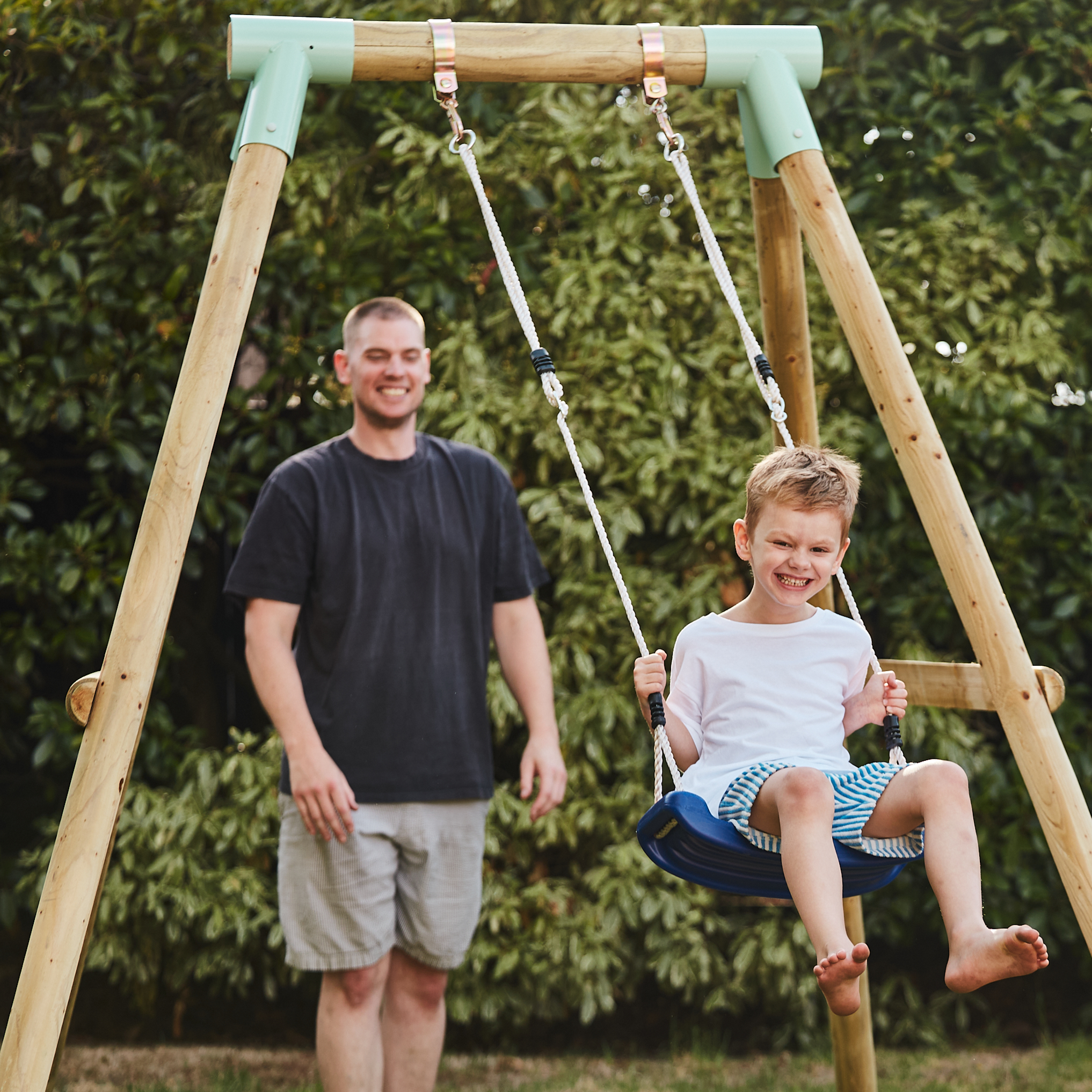 Man standing next to a child on a swing set with greenery in the background